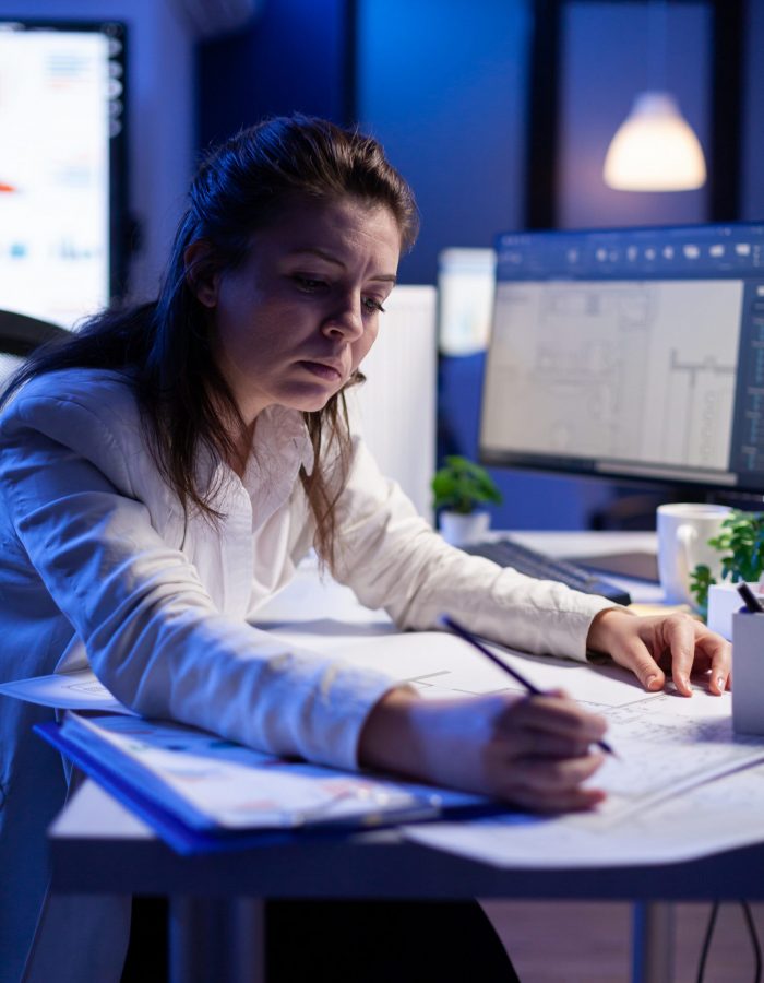 Woman architect analysing and matching blueprints for new building project sitting at desk. Graphic designer using arhitecture prototype plans working overtime in corporate office late at night